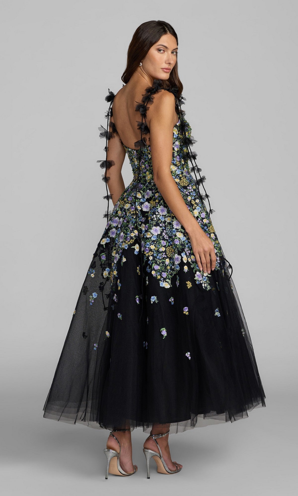 A woman is standing against a gray background, wearing the Mac Duggal 21356 midi party dress—an elegant sleeveless black tulle design featuring an A-line silhouette and multicolored floral embroidery—paired with silver high-heeled sandals.
